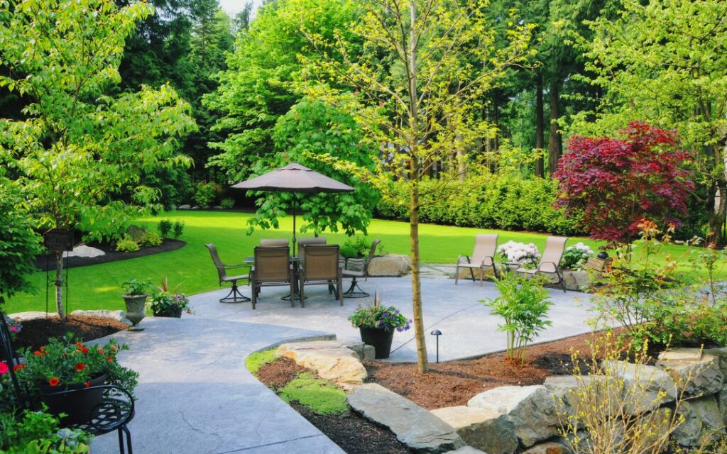 home patio with trees, shrubs and flowers near table, chairs and umbrella