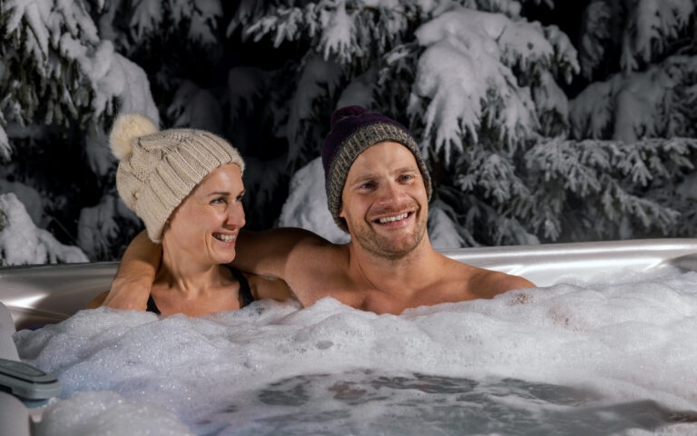 man and woman sitting in a hot tup with stock caps on. Trees with snow behind them.